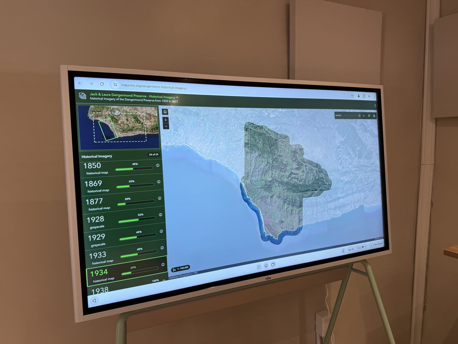 A wall display showing The Nature Conservancy's historical imagery map of the Jack and Laura Dangermond Preserve, its boundary outlined over satellite imagery beside a scrollable list of 29 layers dating from 1850 onward.